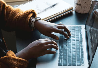 An individual typing on a laptop with a notebook and cup of coffee on the table next to them.